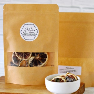 May include: A brown paper bag with a clear window showing dried citrus fruit and lavender. The bag has a white label with the text "Hofoika Home" and a small bird graphic. The bag is sitting on a wooden surface. The text "Rainstorm" is visible on a white label next to the bag. The label also includes the text "Contains: dried sweetgrass, lavender, cloves, rosemary, sage, mint (NOT FOR CONSUMPTION)".