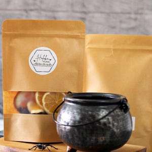 May include: A rustic, black cauldron with three small legs and a handle sits on a wooden board, next to two brown paper bags. One bag has a clear window showing dried orange slices. A small plastic spider adds a Halloween touch.
