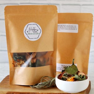 May include: Two brown paper pouches with clear windows, filled with dried botanicals, sit on a wooden board. A small white bowl holds a similar mix. The pouches have a sticker that says "Hofka Home".