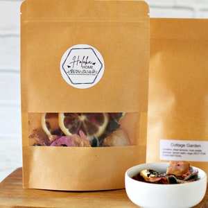 May include: A brown paper bag with a clear window showing dried flowers, citrus fruit, and spices. The bag has a white sticker with the text "Haloka Home" and a floral design. The bag is sitting on a wooden surface next to a white bowl filled with dried flowers and citrus fruit.