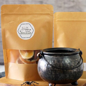 May include: A brown paper bag with a clear window displaying dried orange slices, next to a small black cauldron and a plastic spider on a wooden board. The bag has a white label with the text "Holoka Home".