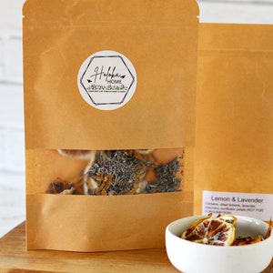 May include: A brown paper bag with a clear window showing dried lemon and lavender inside. The bag has a white label with the text "Hofoka Home" on it. A small white bowl with dried lemon and lavender is on a wooden surface.