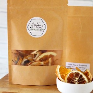 May include: A brown paper bag with a clear window showing dried orange slices and flower petals inside. The bag has a white sticker with the text "Holokai Home" and a small bird graphic. A white bowl with dried orange slices is in the foreground.