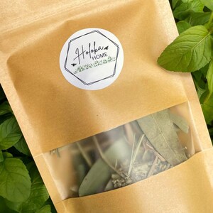 May include: A tan kraft paper pouch with a clear window containing dried botanical leaves and stems. A circular white label on the front features a black geometric frame, the text "Hofoka HOME", and a small illustration of a bee and greenery.