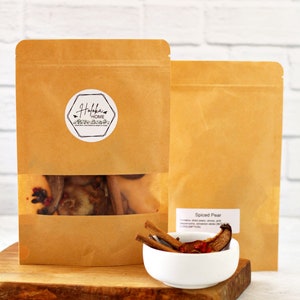 May include: Two brown paper bags, one with a clear window showing dried fruit and spices, and the other plain. A small white bowl holds cinnamon sticks and dried pear slices. The bags have labels with text.