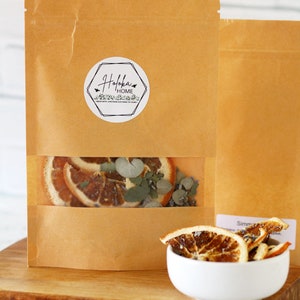 May include: A brown paper bag with a clear window showing dried orange slices, eucalyptus leaves, and lavender. The bag has a white sticker with the text "Hofoka Home" and a small bird graphic. A white bowl with dried orange slices is in the foreground.