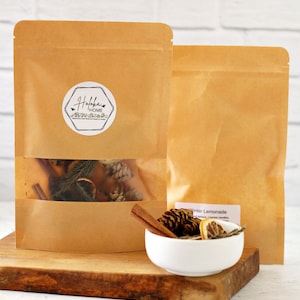 May include: Two brown paper pouches, one with a clear window showing dried botanicals and a logo that says "Hofka Home". A small white bowl contains pinecones, dried citrus, and cinnamon sticks. A small card reads "Winter Lemonade".