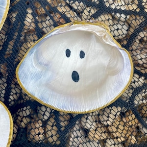 Op de afbeelding: Een witte schelp met een geschilderd spookgezicht, bestaande uit drie zwarte stippen voor ogen en mond. De schelp heeft een goudkleurige rand en ligt op een zwart net met kleine stenen.
