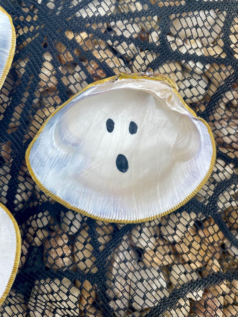 Op de afbeelding: Een witte schelp met een geschilderd spookgezicht, bestaande uit drie zwarte stippen voor ogen en mond. De schelp heeft een goudkleurige rand en ligt op een zwart net met kleine stenen.