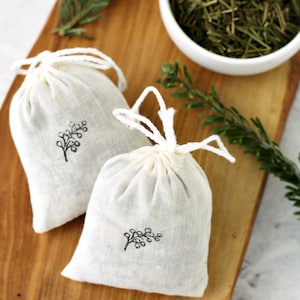 May include: Two white fabric pouches with a sprig of greenery design printed on them. The pouches are tied with white string and are sitting on a wooden cutting board. A bowl of dried herbs is in the background.