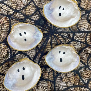 Op de afbeelding: Vier witte spookdecoraties van zeeschelpen met zwarte stipogen en -monden. De schelpen hebben een parelmoerglans en een gouden rand. Ze zijn gerangschikt op een zwart net over een bed van kleine, beige stenen.