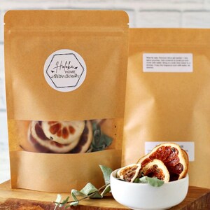 May include: Two brown paper bags with a clear window showing dried orange slices. A small white bowl holds more dried citrus. The bags are on a wooden cutting board. One bag has a sticker that says "Hofoka Home".