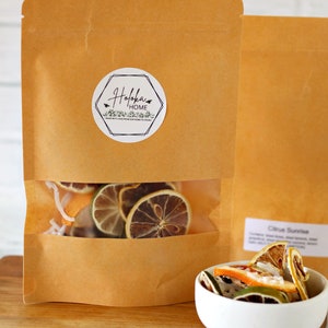 May include: A brown paper bag with a clear window showing dried citrus fruit slices. The bag has a white label with the text "Holokai Home" and a small green leaf design. The bag is sitting on a wooden surface next to a white bowl filled with dried citrus fruit slices.