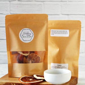 May include: Two brown paper pouches with clear windows, one displaying dried orange slices. A white bowl and dried botanicals sit on a wooden board. The label on the front pouch reads "Haloka Home".
