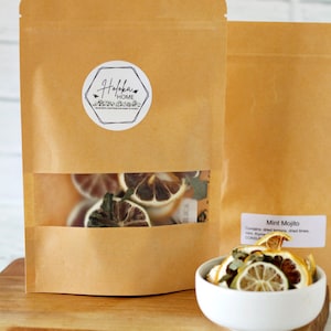 May include: A brown paper bag with a white label that says "Haloka Home" and a clear window showing dried citrus fruit and mint leaves. The bag is sitting on a wooden surface next to a white bowl filled with dried citrus fruit and mint leaves. The bowl is labeled "Mint Mojito" and lists the ingredients: dried lemons, dried limes, mint, thyme, and dried orange.