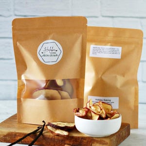 May include: Two brown paper bags with clear windows, one with a label that says "Holiday Home". A white bowl holds dried apple slices. Vanilla beans, cloves, and cinnamon sticks are arranged on a wooden board.