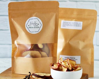 May include: Two brown paper bags with clear windows, one with a label that says "Holiday Home". A white bowl holds dried apple slices. Vanilla beans, cloves, and cinnamon sticks are arranged on a wooden board.
