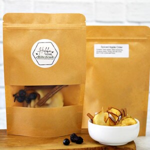 May include: Two brown paper bags with a clear window showing the contents inside. The bag on the left has a white label with the text "Holokai Home" and contains dried fruit, cinnamon sticks, and black peppercorns. The bag on the right has a white label with the text "Spiced Apple Cider". A white bowl with dried apple slices and cinnamon sticks is in front of the bags.
