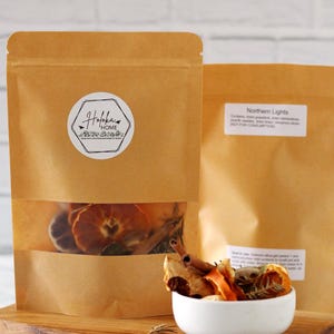 May include: Two brown paper bags with dried fruit and spices. One bag has a clear window showing orange slices and cinnamon sticks. A white bowl holds a portion of the dried ingredients. The bags are on a wooden board.