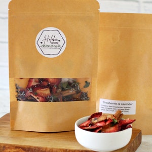 May include: A brown paper bag with a clear window showing dried strawberries, lavender, and cinnamon sticks. The bag has a white label with the text "Holokau HOME" on it. The bag is sitting on a wooden surface next to a white bowl filled with dried strawberries and lavender. The text "Strawberries & Lavender" is visible on the bag, along with the text "Contains dried strawberries, lavender, mint, cinnamon stick, nemesia flower petals (NOT FOR CONSUMPTION)"
