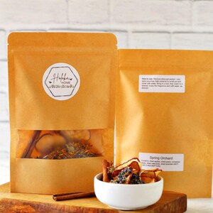 May include: Two brown paper pouches with a clear window, filled with dried herbs and spices. A white bowl holds dried fruit and lavender. Cinnamon sticks are on a wooden cutting board. The label on the pouch reads "Hafkba Home".