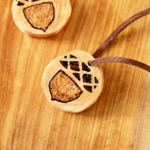 May include: Two wooden acorn pendants with brown leather cords. The acorns are burned with a dark brown design.