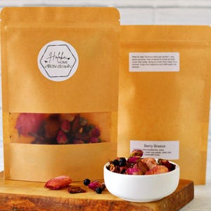 May include: Two brown paper bags with a white label that says "Holokai Home" and a white circle with a small bird design. The bag on the left has a clear window showing dried rose petals and berries. The bag on the right has a white label that says "Berry Breeze" and a description of the product. A white bowl filled with dried rose petals and berries sits on a wooden surface.