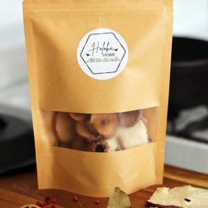May include: A tan kraft paper stand-up pouch with a clear window containing dried fruit slices. A circular white label features the text "Hofoka HOME" inside a black hexagonal outline with small bee illustrations and green foliage graphics.