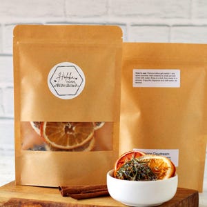 May include: Two brown paper bags with clear windows showing dried orange slices and lavender. The bags are labeled "Halfbak HOME". A white bowl with dried orange slices and lavender is on a wooden surface. Cinnamon sticks are on the surface.