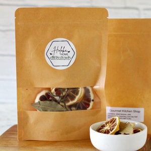 May include: A brown paper bag with a white label that says "Holokai Home" and a clear window showing dried citrus fruit, bay leaves, and other spices. The bag is sitting on a wooden cutting board next to a white bowl with dried citrus fruit and bay leaves.