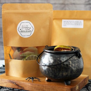 May include: A black cauldron with dried citrus fruit inside, sitting on a wooden board with a black spider. Two brown paper bags with the text "Holokai Home" are in the background.