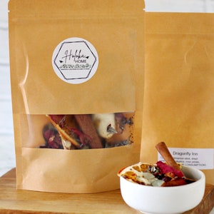 May include: Two brown paper pouches with a clear window displaying dried botanicals, including cinnamon sticks and rose petals. A small white bowl holds a similar mix, topped with a cinnamon stick. The pouches have a circular label that says "Hofka Home".