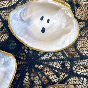 Op de afbeelding: Een close-up van een witte zeeschelp met een goud geschilderde rand. De binnenkant van de schelp heeft drie zwarte stippen geschilderd om op een spookgezicht te lijken. De schelp rust op een zwart net.