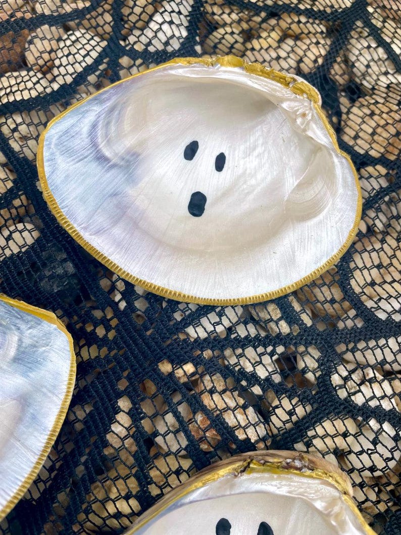 Op de afbeelding: Een close-up van een witte zeeschelp met een goud geschilderde rand. De binnenkant van de schelp heeft drie zwarte stippen geschilderd om op een spookgezicht te lijken. De schelp rust op een zwart net.