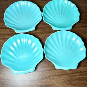 May include: Four turquoise seashell-shaped dishes. Each dish has a scalloped edge and a fan-like pattern radiating from the center. The dishes are made of a solid, opaque material and are arranged on a wooden surface.