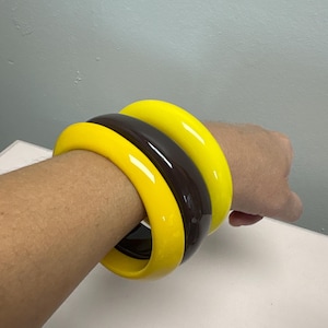 May include: A set of three chunky bangle bracelets featuring a smooth, glossy finish. The set includes two vibrant yellow bangles and one dark brown bangle, all with a rounded, thick profile and a solid, uniform color throughout.