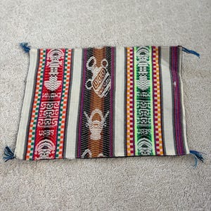 May include: Rectangular woven textile with a cream background and vertical stripes in red, orange, green, and brown. The design includes geometric patterns and stylized figures. Blue tassels are on each corner.