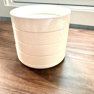 May include: A cylindrical off-white ceramic planter featuring three horizontal recessed grooves encircling the exterior. The vessel has a smooth, glossy finish and a wide, open top, presenting a clean, uniform appearance throughout the entire structure.