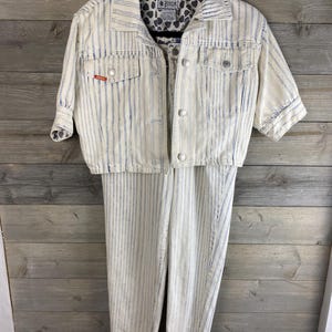 May include: A vintage two-piece denim set with vertical blue stripes on a cream background. The set includes a short-sleeved jacket with button closures and a pair of matching pants. The jacket has two front pockets and a patterned lining.