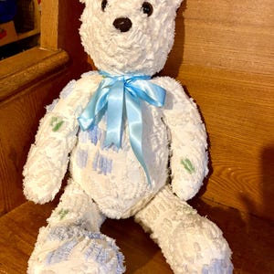 May include: A white teddy bear with a light blue satin ribbon tied around its neck. The bear has black button eyes and a dark brown nose. Patches of blue and green fabric are sewn onto the bear's body.