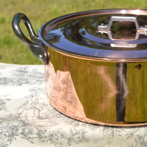 以下が含まれることがあります： 黒い取っ手と銀色の取っ手のある銅製の蓋が付いた銅製の鍋。鍋は花柄の白いテーブルクロスの上に置かれています。