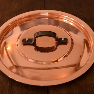 Peut inclure: Un couvercle de casserole en cuivre brillant avec une poignée en métal noir. Le couvercle a une surface lisse et réfléchissante.