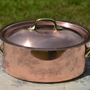 May include: A round copper cooking pot with a matching lid and two brass side handles. The pot features a polished metallic finish, a smooth cylindrical body, and a domed lid with a central brass handle, reflecting light across its surface.