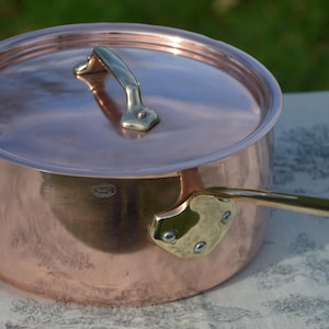 May include: A copper cooking pot with a lid and a long handle. The pot has a shiny, reflective surface and a brass handle. The lid also has a brass handle. The pot is sitting on a patterned surface.