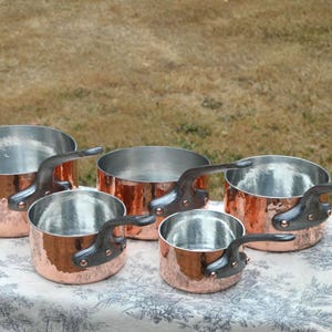 Pode incluir: Um conjunto de quatro panelas redondas de cobre com interiores prateados e cabos metálicos escuros e curvos fixados com rebites de cobre. As panelas variam em tamanho e apresentam um acabamento exterior polido e brilhante.