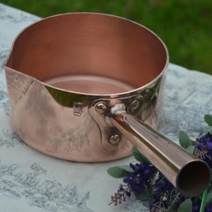 May include: A copper saucepan with a long handle. The pan has a rounded shape with a pouring spout. The handle is attached with rivets. The pan is sitting on a patterned surface with purple flowers.
