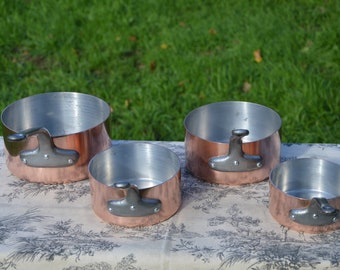 May include: Four copper cooking pots with silver interiors and riveted handles are arranged on a patterned tablecloth. The pots vary in size, with a bright copper exterior and a shiny, reflective interior. The background is a blurred green lawn.