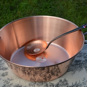 Op de afbeelding: Een grote koperen pan met zwarte handvatten en een koperen schep met een geperforeerde kom. De pan heeft een decoratief ontwerp op de bodem.