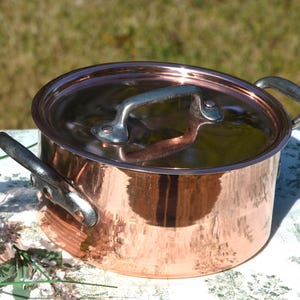 Puede incluir: Una olla redonda de cobre con tapa a juego y dos asas laterales. La olla presenta un acabado metálico pulido y un asa central en la tapa. La construcción incluye asas de metal plateado fijadas con remaches a los lados y a la tapa.