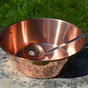 May include: A large copper pot with two handles and a copper ladle with holes. The pot has a decorative design on the bottom and is sitting on a white tablecloth with a floral pattern.
